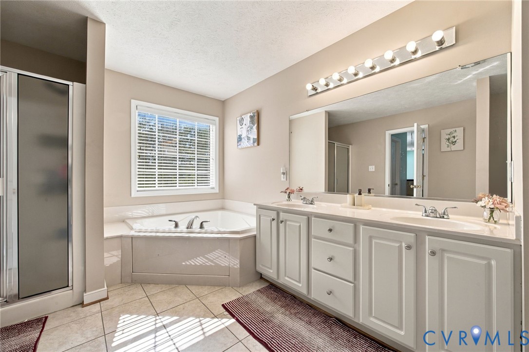 Primary bathroom with Dual-sink vanity with white cabinetry, expansive wall-mounted mirror, corner soaking tub, and separate glass-enclosed shower