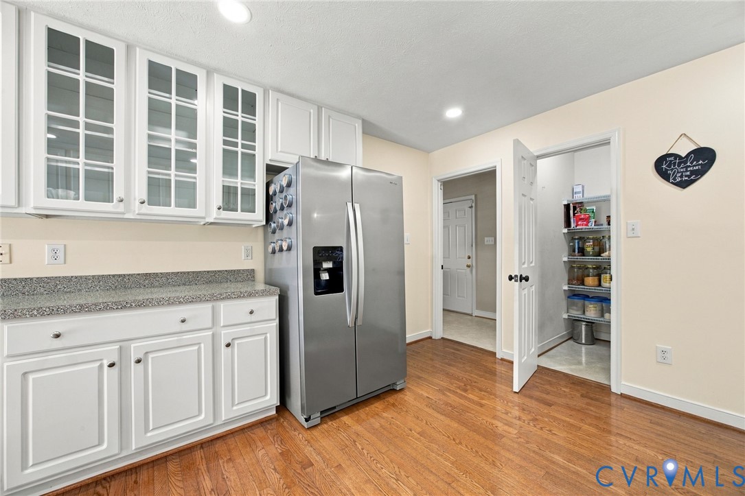 Kitchen featuring white cabinetry with glass-front uppers, speckled stone-finish countertops, stainless steel side-by-side refrigerator, wood-finish flooring, and a walk-in pantry Kitchen featuring white cabinetry with glass-front uppers, speckled stone-finish countertops, stainless steel side-by-side refrigerator, wood-finish flooring, and a walk-in pantry