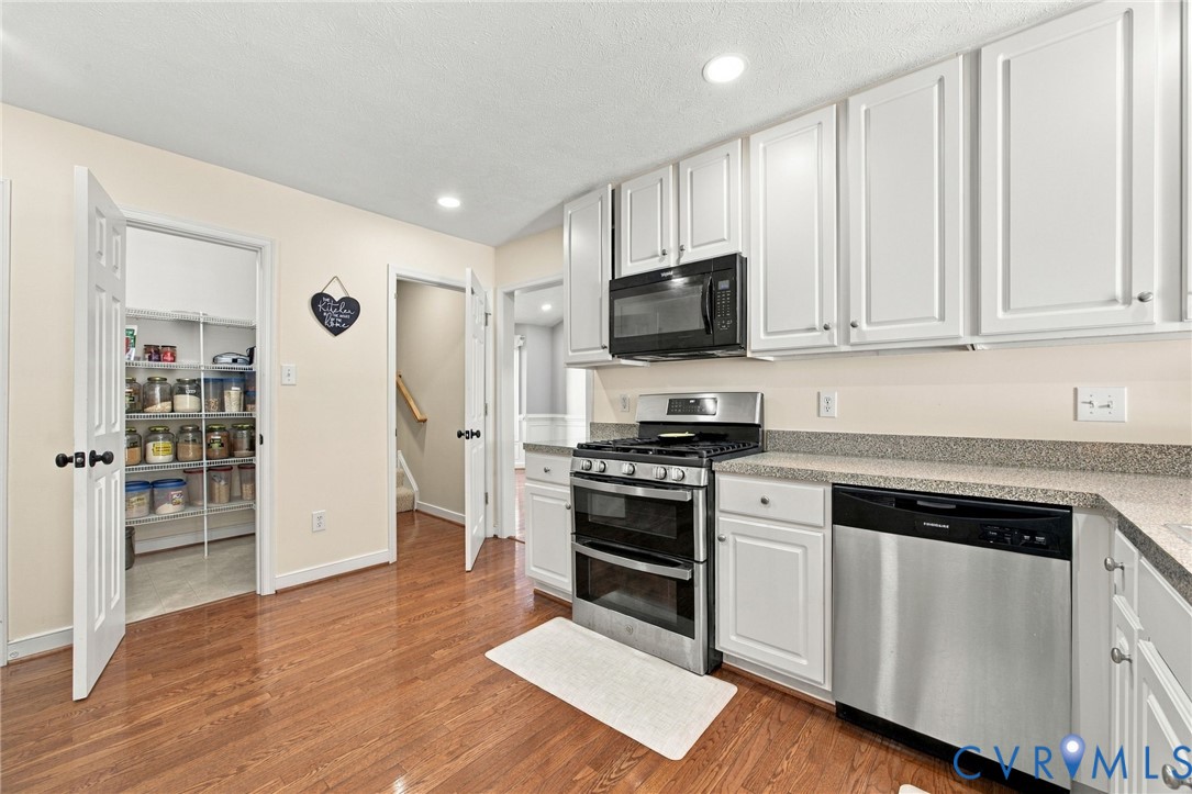 Kitchen featuring white shaker-style cabinetry, wood-finish flooring, stone-composite countertops, stainless steel appliances, and a walk-in pantry Kitchen featuring white shaker-style cabinetry, wood-finish flooring, stone-composite countertops, stainless steel appliances, and a walk-in pantry