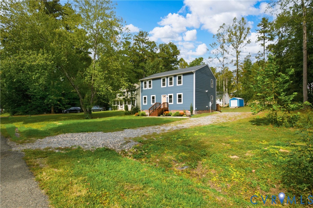 Two-story residence featuring blue horizontal siding, a gravel driveway, an elevated front wooden deck, and a detached storage structure on a tree-lined lot