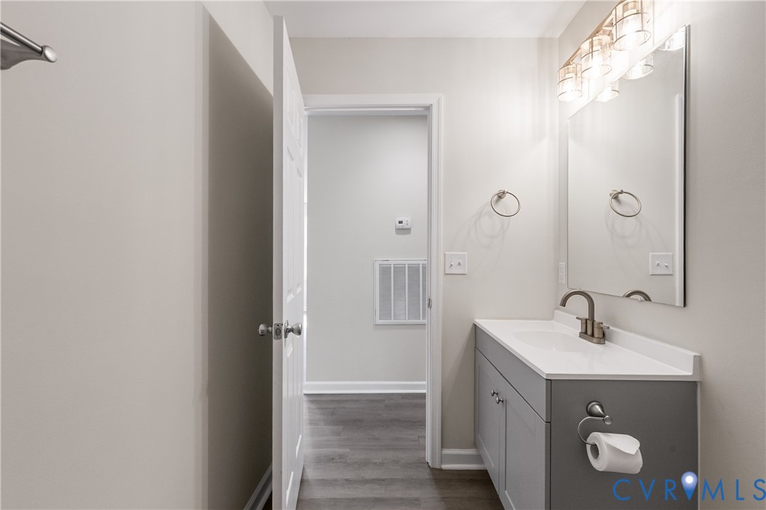 Grey vanity featuring a solid surface countertop, integrated sink, and brushed-finish faucet, accompanied by a rectangular wall-mounted mirror, multi-bulb vanity light fixture, and wood-finish flooring