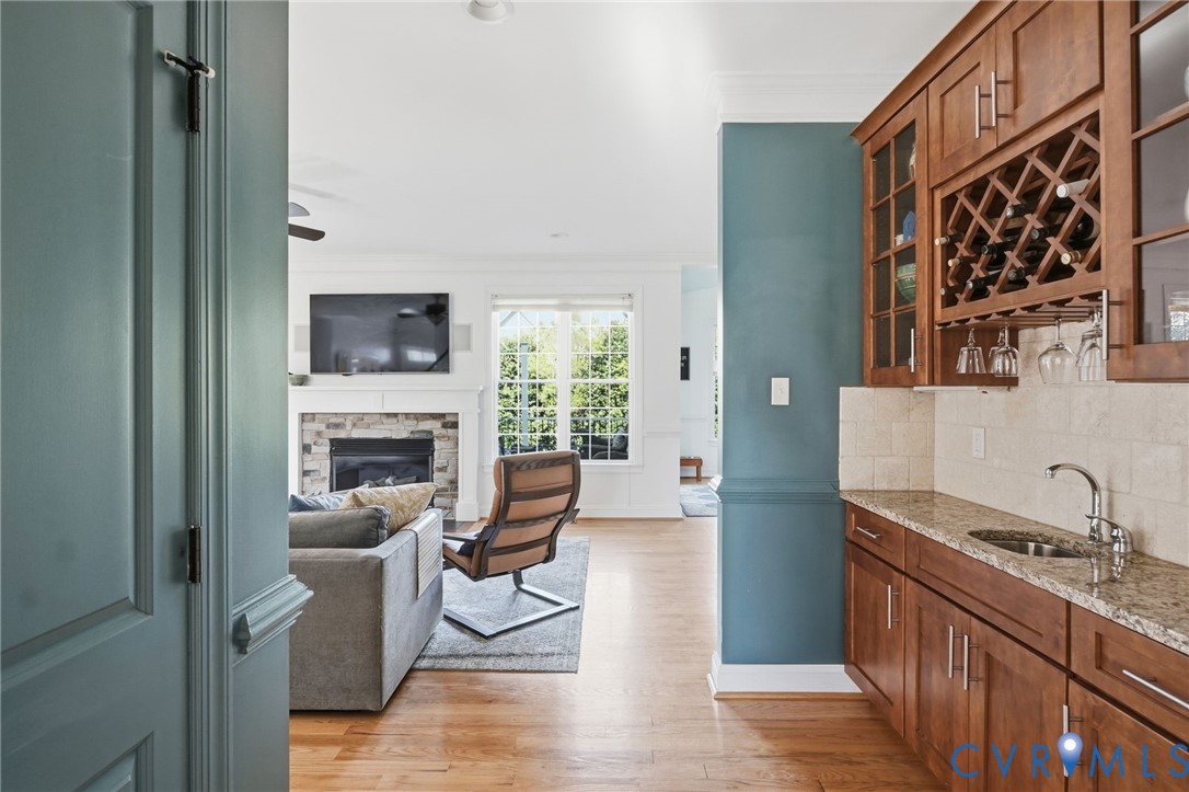 Built-in wet bar featuring stone-surface countertops, custom wood cabinetry, integrated wine storage, and a stainless steel prep sink, complemented by wood-finish flooring and a fireplace with a stone-surround mantel