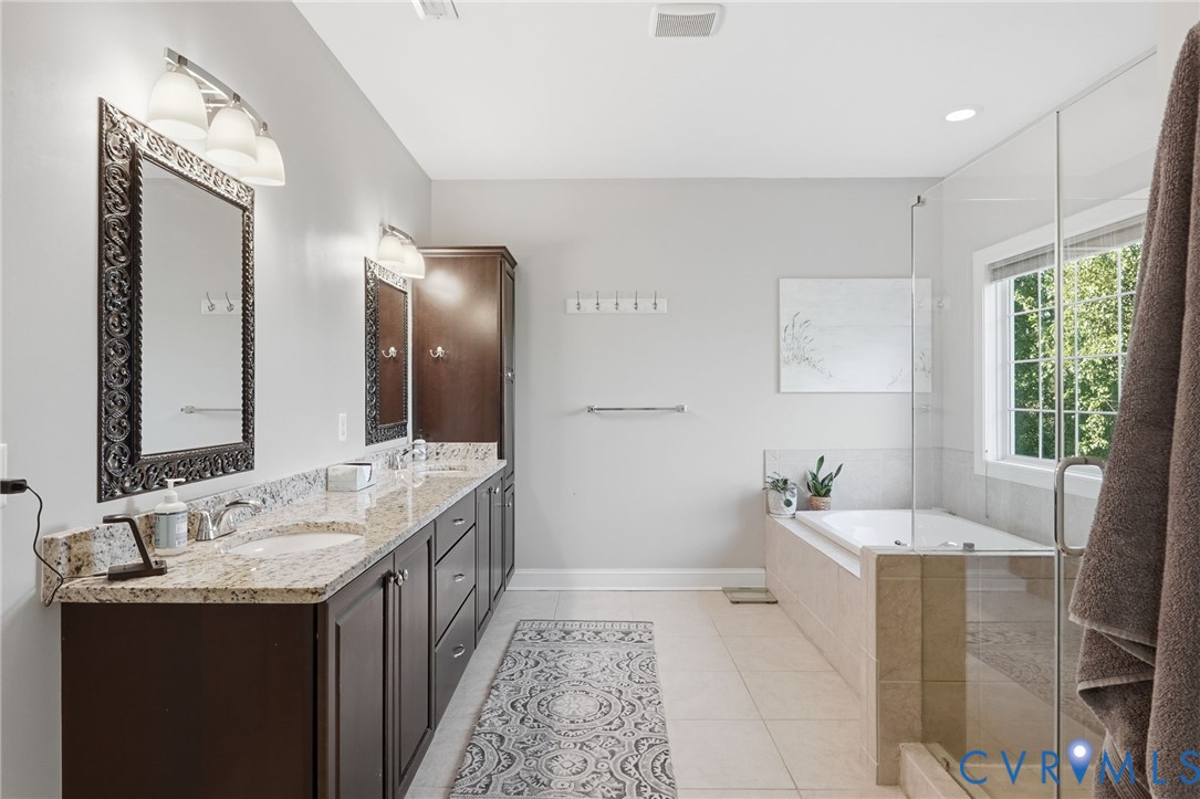 Double vanity with stone countertop, dark wood-finish cabinetry, tile flooring, glass-enclosed shower, and a built-in soaking tub