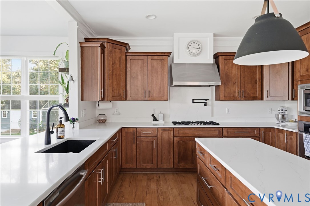 Kitchen featuring stained wood cabinetry, stone countertops, a stainless steel gas range with a matching wall-mounted hood, and an oversized black pendant light fixture Kitchen featuring stained wood cabinetry, stone countertops, a stainless steel gas range with a matching wall-mounted hood, and an oversized black pendant light fixture