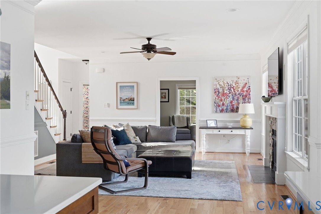 Living area featuring wood-finish flooring, a fireplace with a stone-surround hearth, crown molding, chair rail trim, and a ceiling fan Living area featuring wood-finish flooring, a fireplace with a stone-surround hearth, crown molding, chair rail trim, and a ceiling fan
