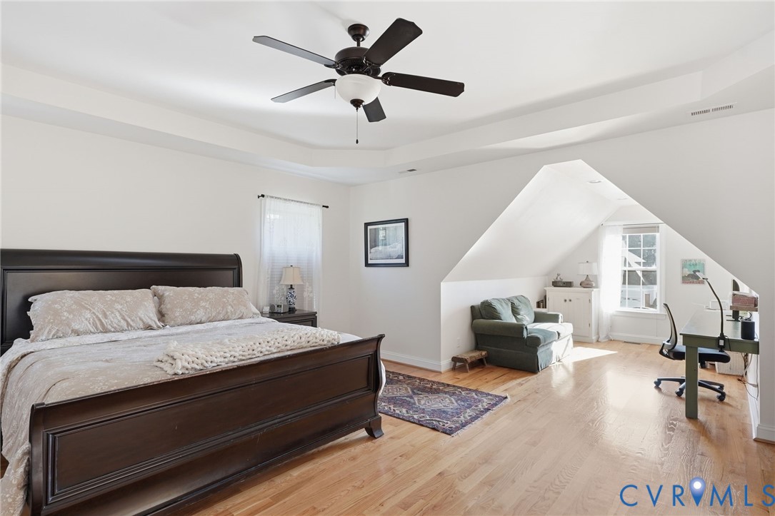Tray ceiling, wood-finish flooring, and multi-paned windows featuring architectural dormer alcoves and an attached ceiling fan with integrated lighting