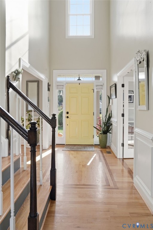 Entryway featuring a two-story ceiling with high-elevation window, wood-finish flooring with inlaid border, staircase with dark wood-finish railing, and a paneled door with glass side-lights