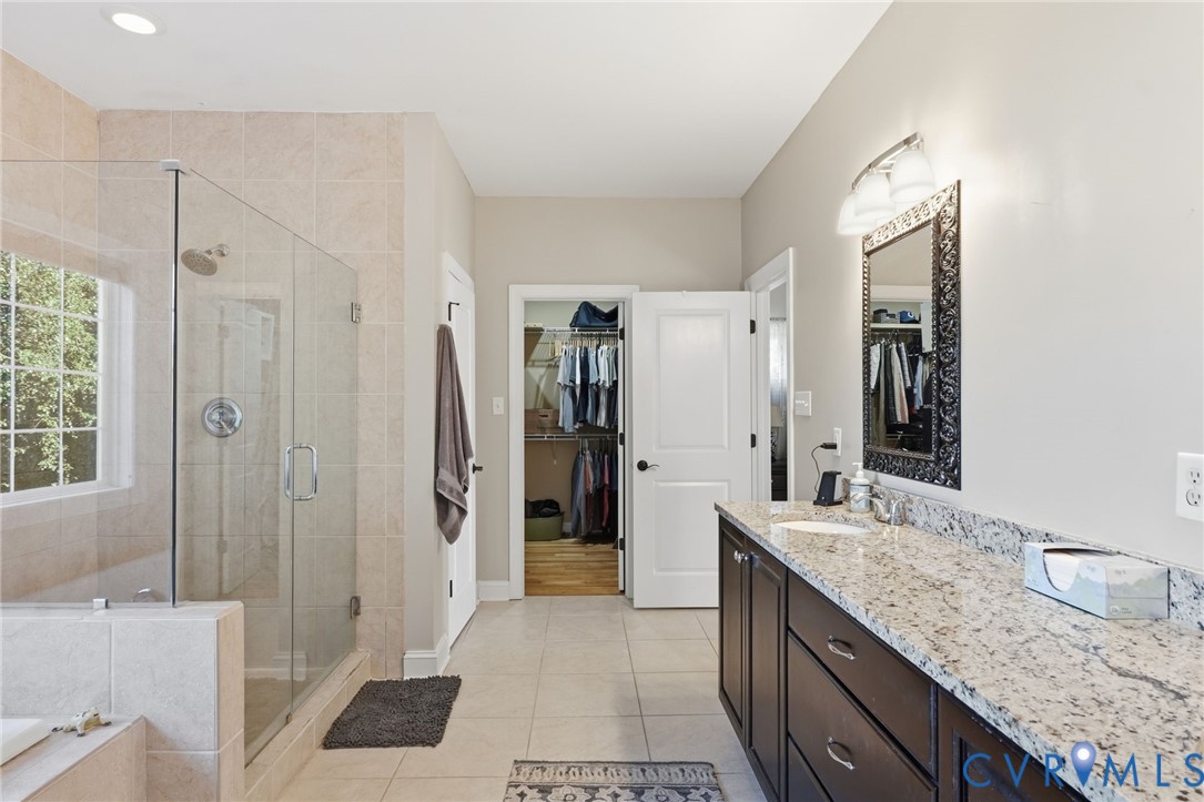 Bathroom featuring a glass-enclosed shower, stone-surfaced vanity with dual sinks, dark-finish cabinetry, and ceramic tile flooring