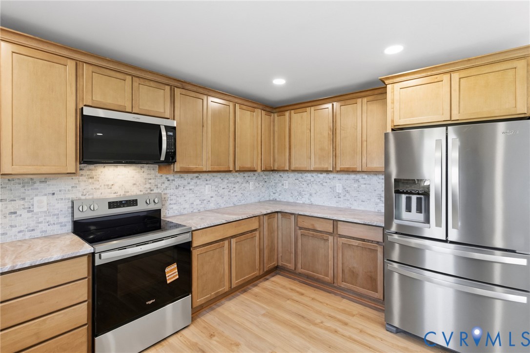 Kitchen featuring light wood-finish cabinetry, stone-patterned countertops, a multi-tone tile backsplash, and stainless steel appliances