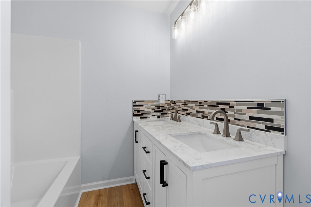 Bathroom vanity featuring a light-toned stone-finish countertop, dual under-mount sinks, linear multi-colored mosaic tile backsplash, matte black hardware, and wood-finish flooring