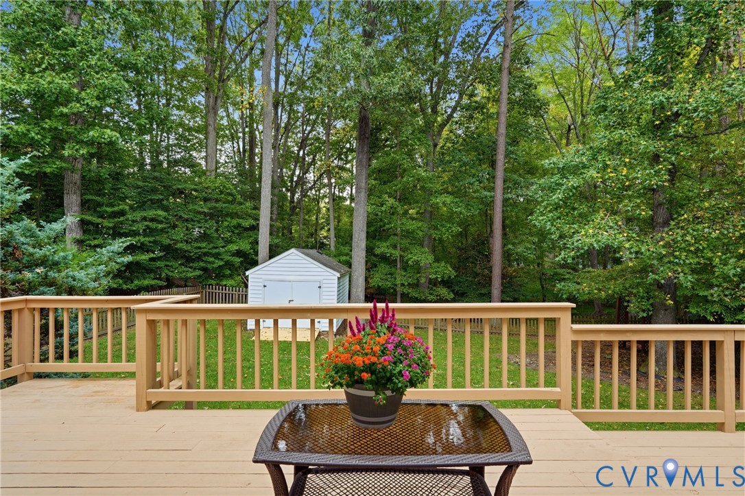 Raised wood deck with balustrades, expansive lawn, detached storage structure, and perimeter fencing set against a wooded backdrop
