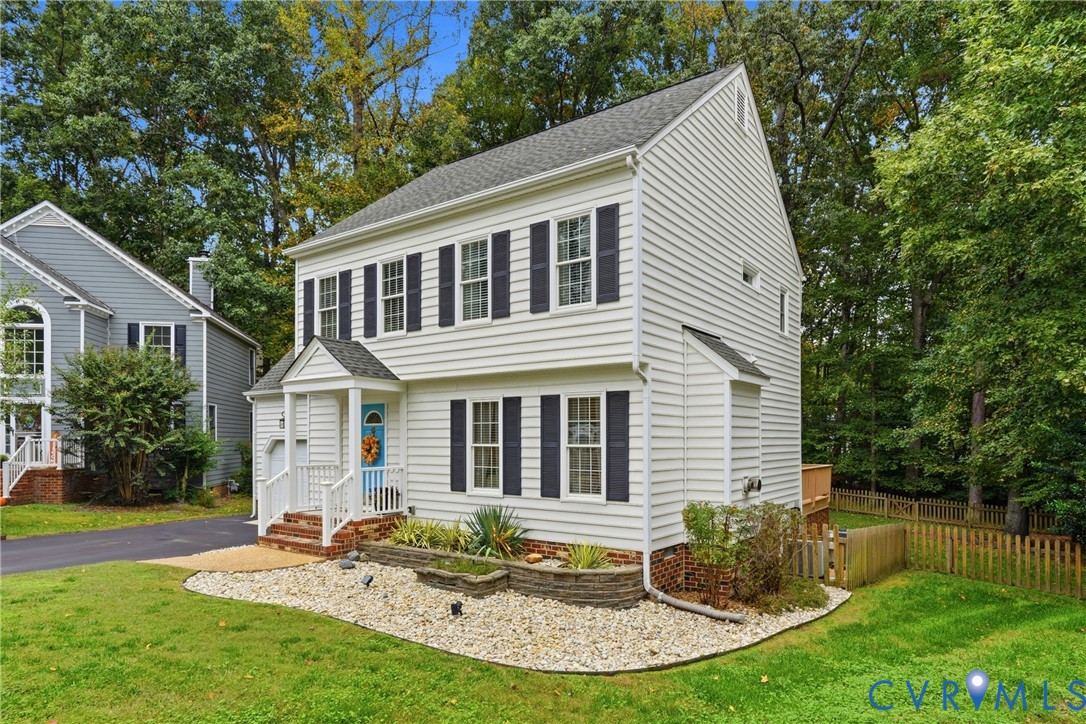 Two-story colonial exterior with white horizontal siding, dark shutters, covered front porch, brick-accented foundation, and tiered stone landscape retaining walls