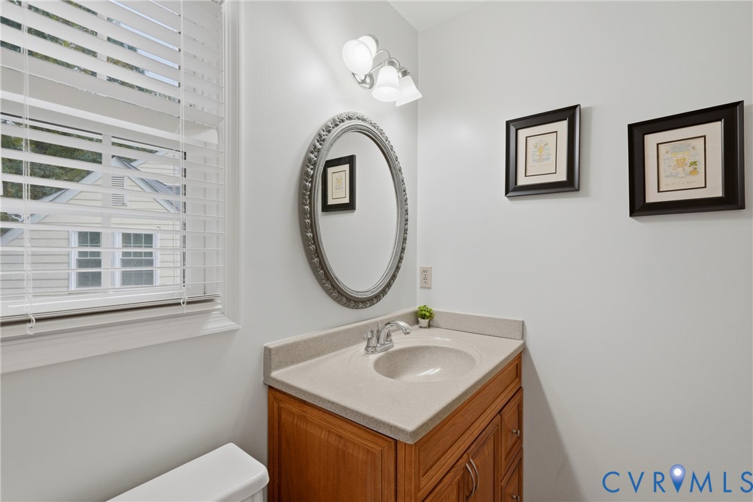 Wooden vanity featuring a stone-composite countertop with an integrated basin, oval wall-mounted mirror, three-light fixture, and horizontal blind window treatment