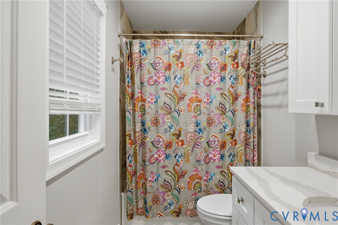 Bathroom featuring a white shaker-style vanity with a veined stone countertop, tub with neutral tiled surround, white horizontal blinds, and wall-mounted metal shelving