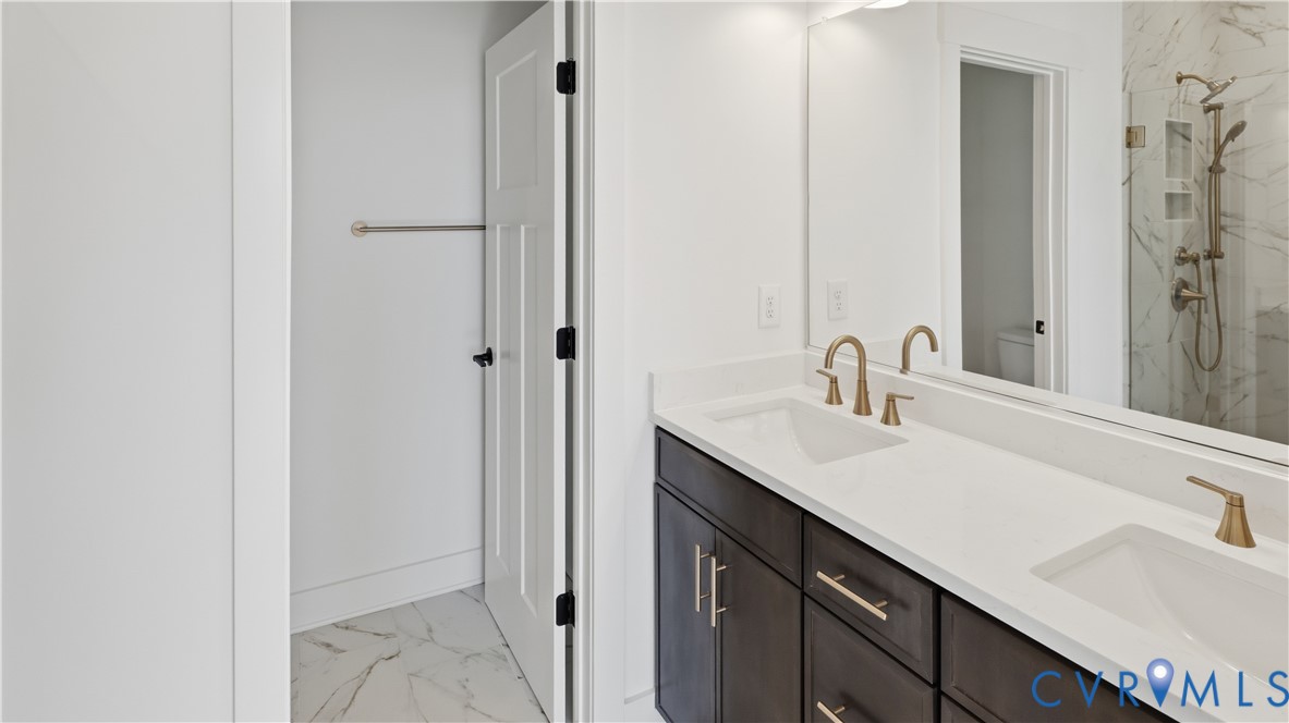 Double vanity featuring dark cabinetry, quartz countertops, dual under-mount sinks with brass-tone fixtures, and a walk-in shower with stone-finish tile surround