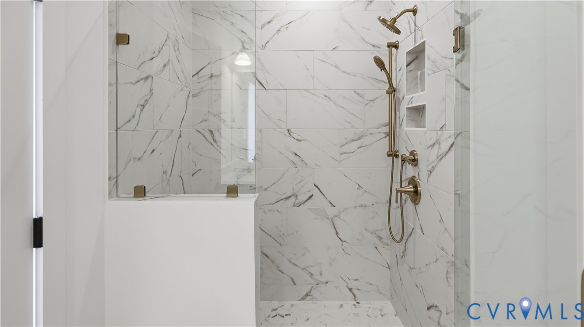Walk-in shower featuring large-format stone-patterned tile, frameless glass enclosure, built-in recessed wall shelving, and gold-tone hardware including a fixed rain-style showerhead and handheld sprayer