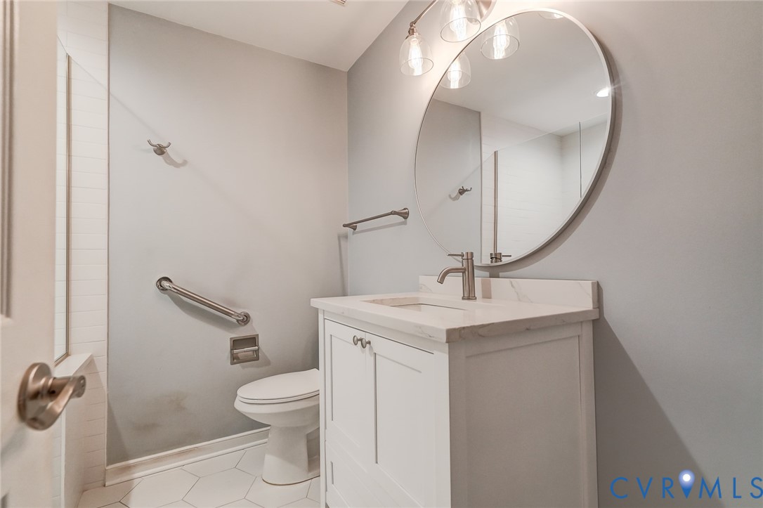 Bathroom featuring a white vanity cabinet with a stone-top integrated sink, round frameless mirror, three-light fixture, and hexagonal tile flooring