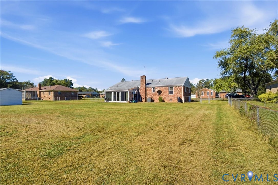 Expansive backyard featuring a large grass lawn, chain-link perimeter fencing, brick exterior construction, rear-facing chimney, and glass-enclosed porch