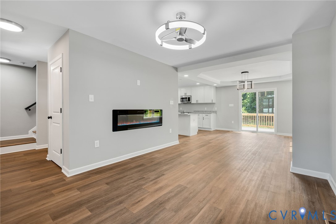 Open-concept interior featuring wood-finish flooring, a wall-mounted electric fireplace, modern circular flush-mount lighting, and sliding glass doors with access to an exterior deck Open-concept interior featuring wood-finish flooring, a wall-mounted electric fireplace, modern circular flush-mount lighting, and sliding glass doors with access to an exterior deck