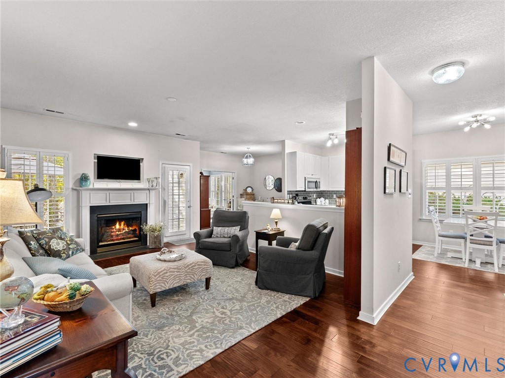 Open living area featuring wood-finish flooring, a fireplace with a white mantel, white cabinetry, and plantation shutters