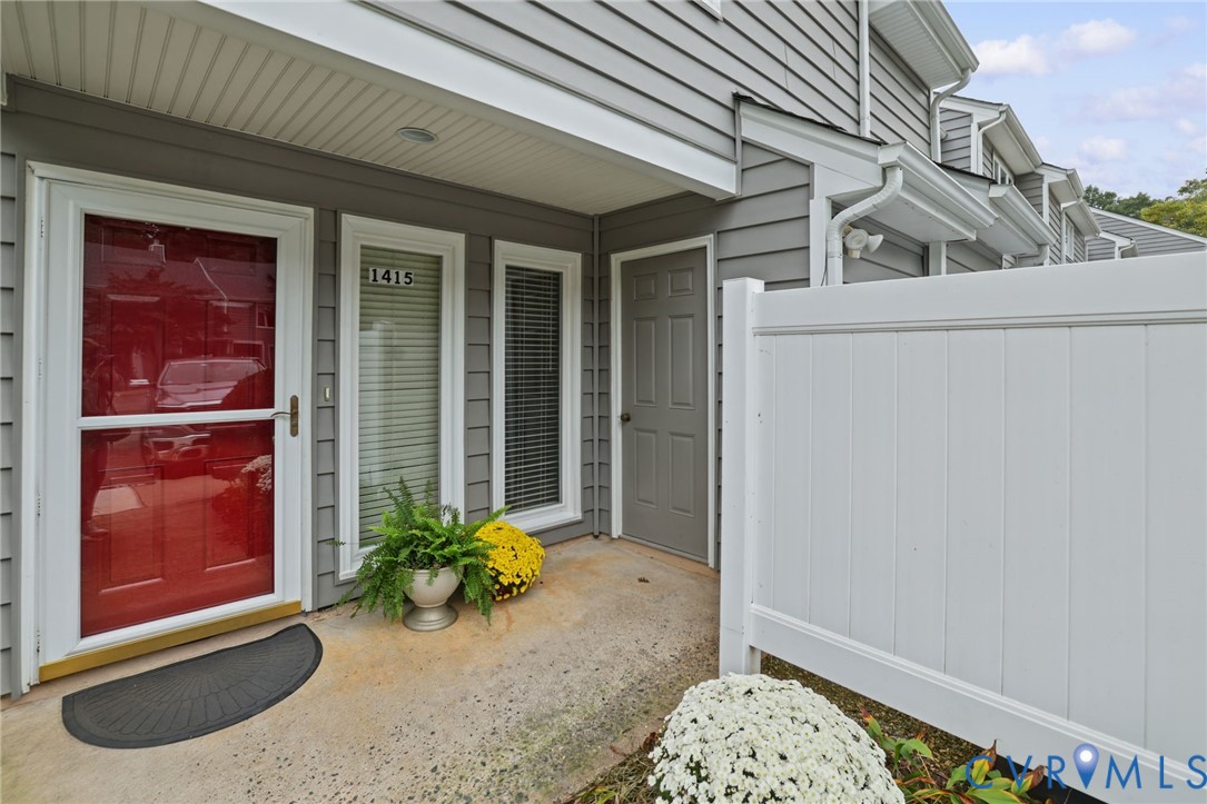 Covered entryway featuring gray horizontal lap siding, a red front door with a glass storm door, dual vertical windows, and a white privacy fence