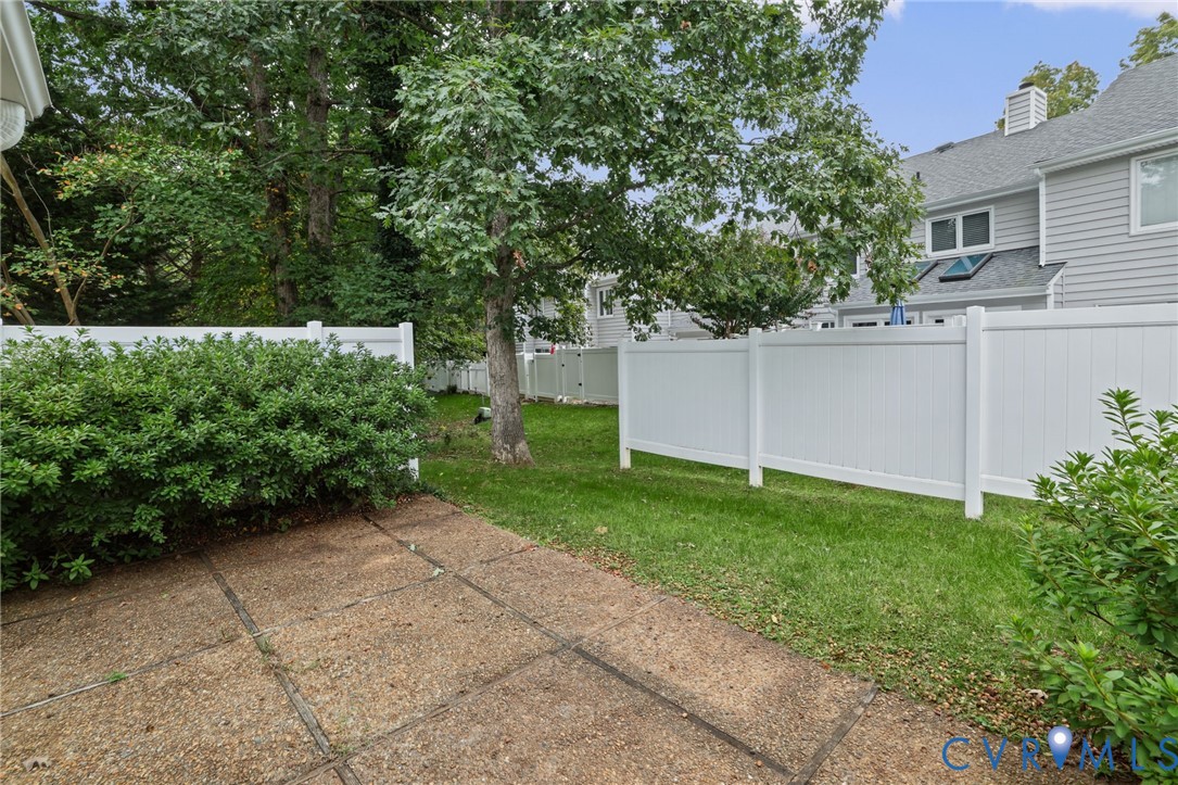 Private rear patio featuring aggregate concrete flooring, enclosed by a white vinyl privacy fence and bordered by mature trees and manicured landscaping