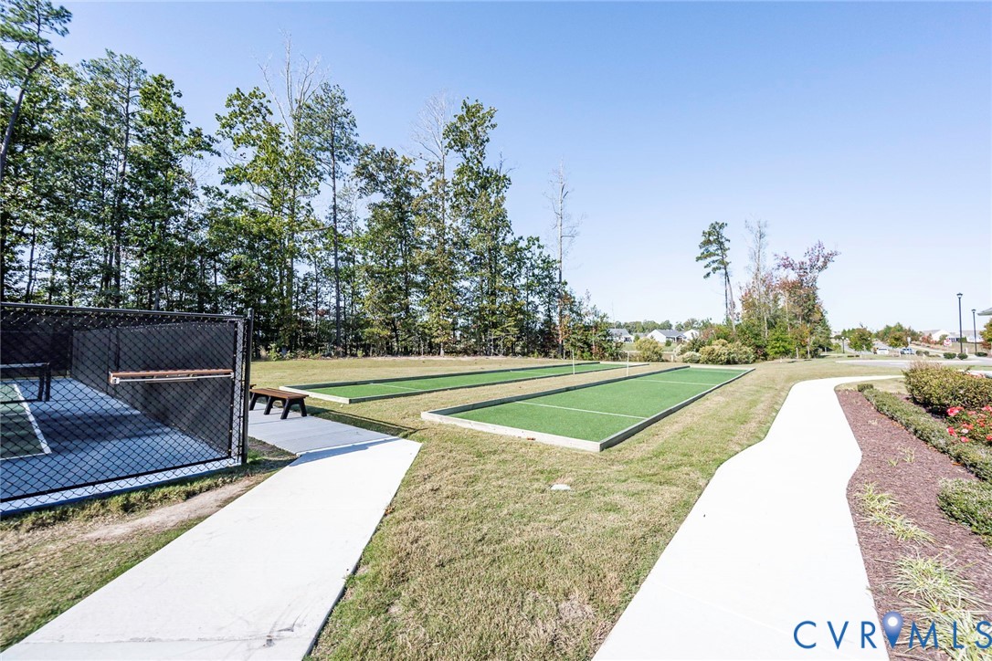 Outdoor recreation area featuring two bocce ball courts with synthetic turf, perimeter chain-link fencing, concrete walking paths, and mature woodland borders
