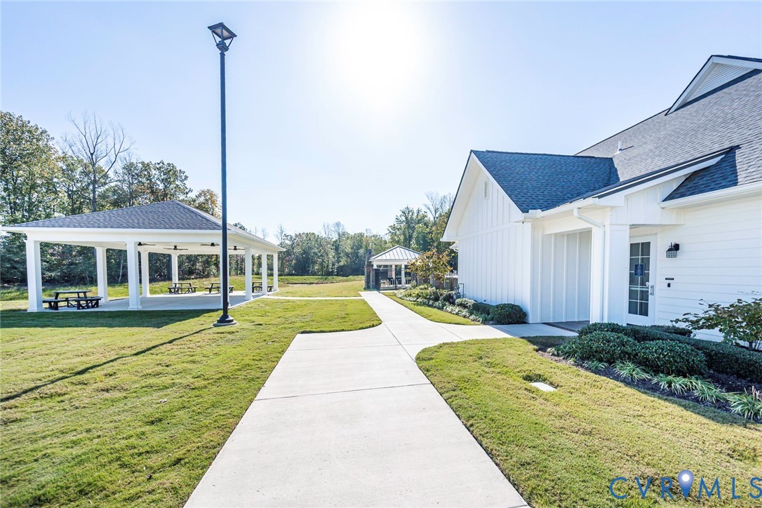 White board-and-batten siding with a dark asphalt shingle roof, concrete walkways, expansive manicured lawn, and a covered open-air pavilion