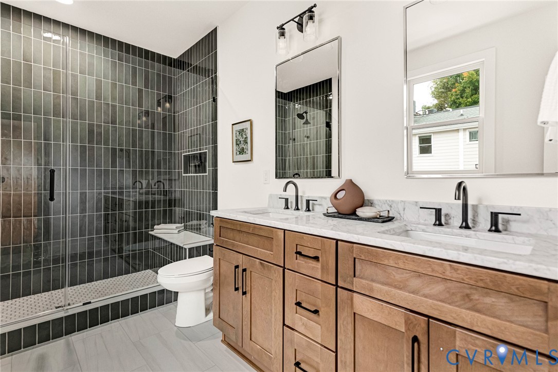 Double vanity with stone countertop and undermount sinks, wood-finish cabinetry, matte black plumbing fixtures, walk-in shower with vertical tile surround and integrated bench, and rectangular wall-mounted mirrors