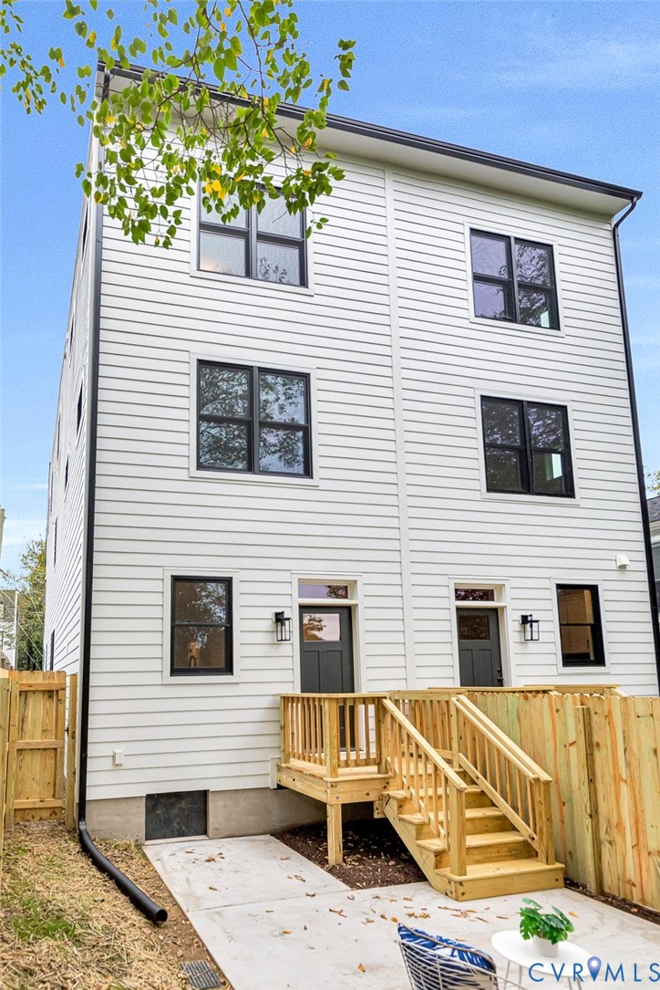 Three-story horizontal lap siding exterior with black-framed windows, two rear entry doors, wall-mounted exterior lanterns, and a wood deck with stairs leading to a concrete patio