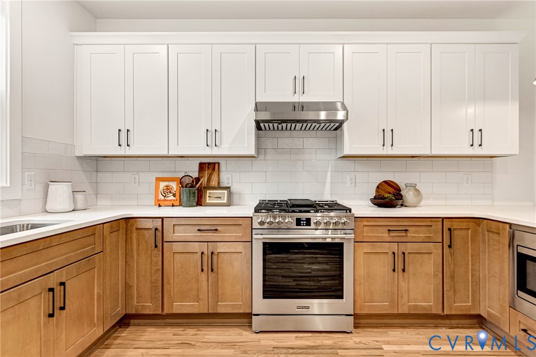 Kitchen featuring two-tone cabinetry with white upper and light-toned lower units, subway tile backsplash, stainless steel gas range, stainless steel range hood, and solid-surface countertops