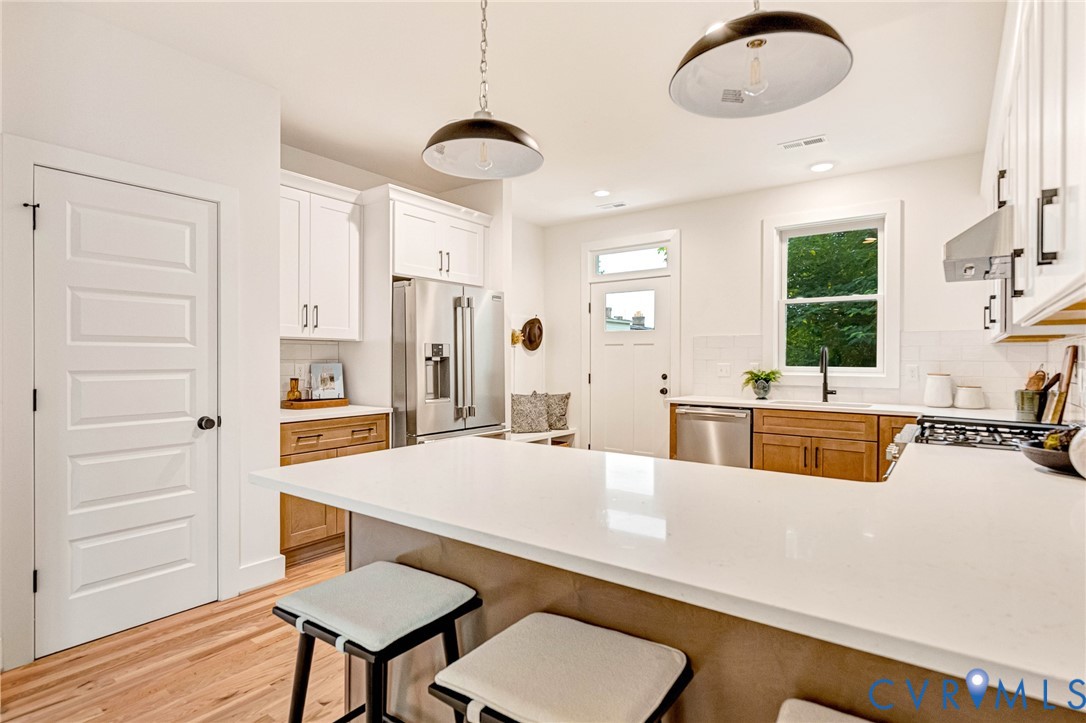 Kitchen featuring white stone countertops, a combination of white upper and natural wood lower cabinetry, stainless steel appliances, wood-finish flooring, and industrial-style pendant lighting