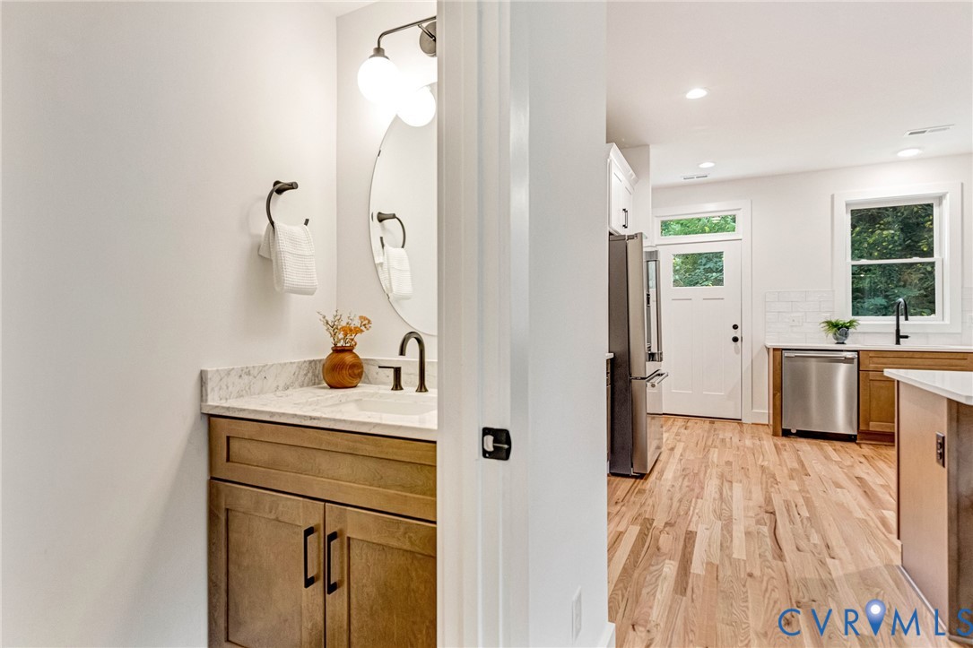 Half-bath featuring a vanity with stone-finish countertop, integrated sink, and circular mirror, along with wood-finish flooring extending into the kitchen area