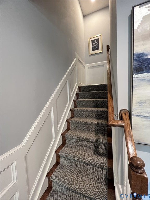 Staircase featuring white wainscoting, wooden handrail with a decorative newel post, and patterned carpet runner over wood-finish treads Staircase featuring white wainscoting, wooden handrail with a decorative newel post, and patterned carpet runner over wood-finish treads