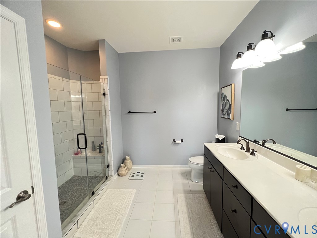 Bathroom featuring a dual-sink vanity with a solid-surface countertop, dark cabinetry, a glass-enclosed shower with subway tile surround, and a large wall-mounted mirror with integrated lighting