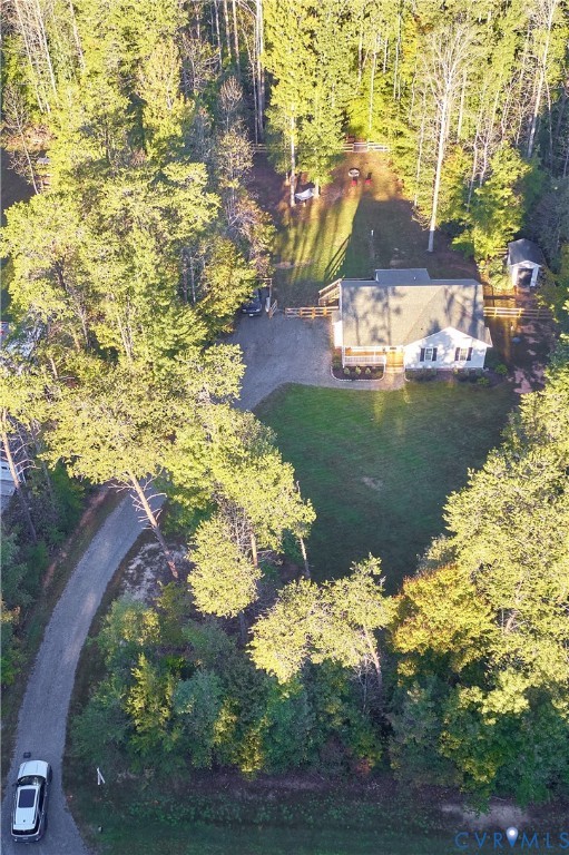 Aerial view of a residential property featuring a gravel driveway, expansive lawn, detached storage structure, and perimeter fencing, all situated within a mature wooded setting