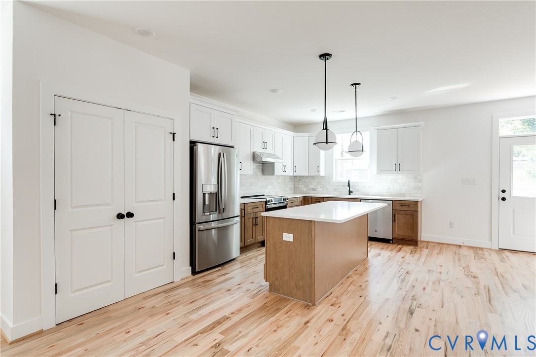 Kitchen featuring white upper cabinetry, lower wood-finish cabinetry, an island with a light-colored countertop, stainless steel appliances, and wood-finish flooring Kitchen featuring white upper cabinetry, lower wood-finish cabinetry, an island with a light-colored countertop, stainless steel appliances, and wood-finish flooring