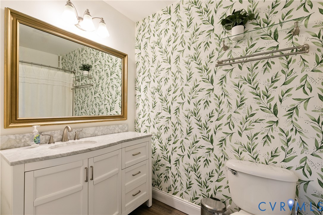 White vanity with stone-finish countertop, integrated sink, gold-tone framed mirror, three-light fixture, and wall-mounted metal shelving