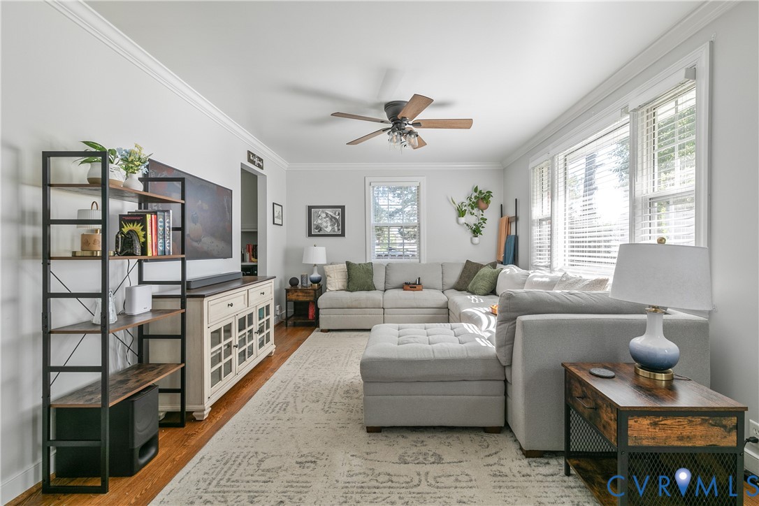 Living room featuring wood-finish flooring, crown molding, a central ceiling fan, and multi-pane windows