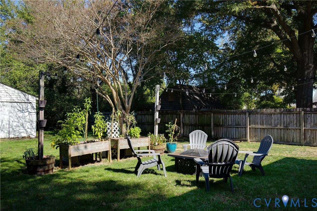 Spacious backyard featuring a wood-panel fence, raised wooden garden beds, string lighting, and mature trees