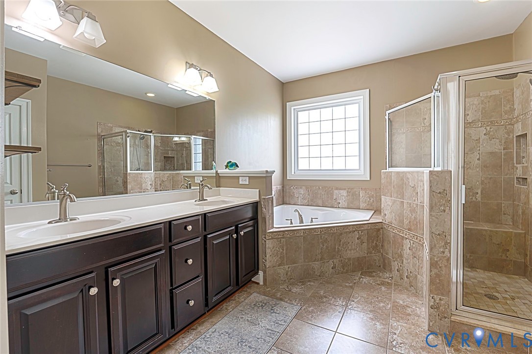 Dual-sink vanity with dark-finish cabinetry, solid-surface countertops, corner soaking tub, and enclosed glass-door shower with stone-tile surround