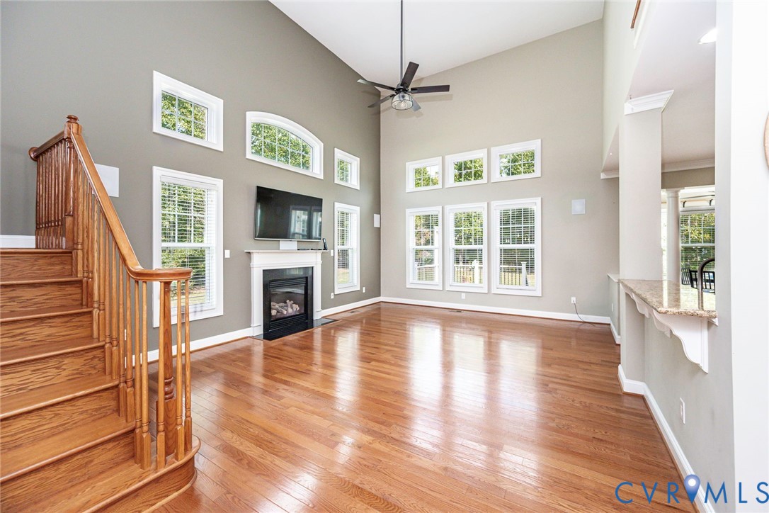 Vaulted ceilings featuring a central fan, hardwood flooring, multi-level windows, a gas fireplace with a decorative mantel, and a wooden staircase with a baluster railing