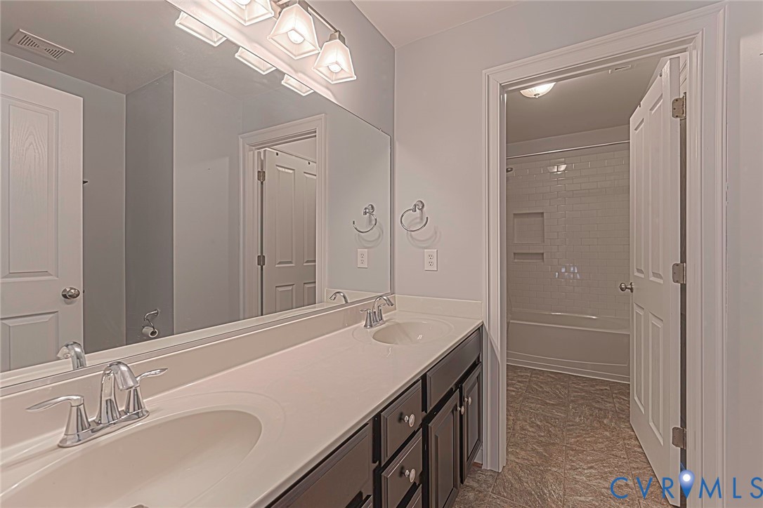 Dual-sink vanity with a solid-surface countertop, full-width mirror, multi-light fixture, and recessed tub-shower combination with tile surround