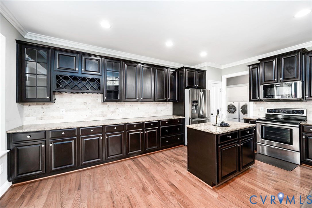 Kitchen featuring dark cabinetry with built-in wine storage, stone countertops, stone tile backsplash, stainless steel appliances, and wood-finish flooring