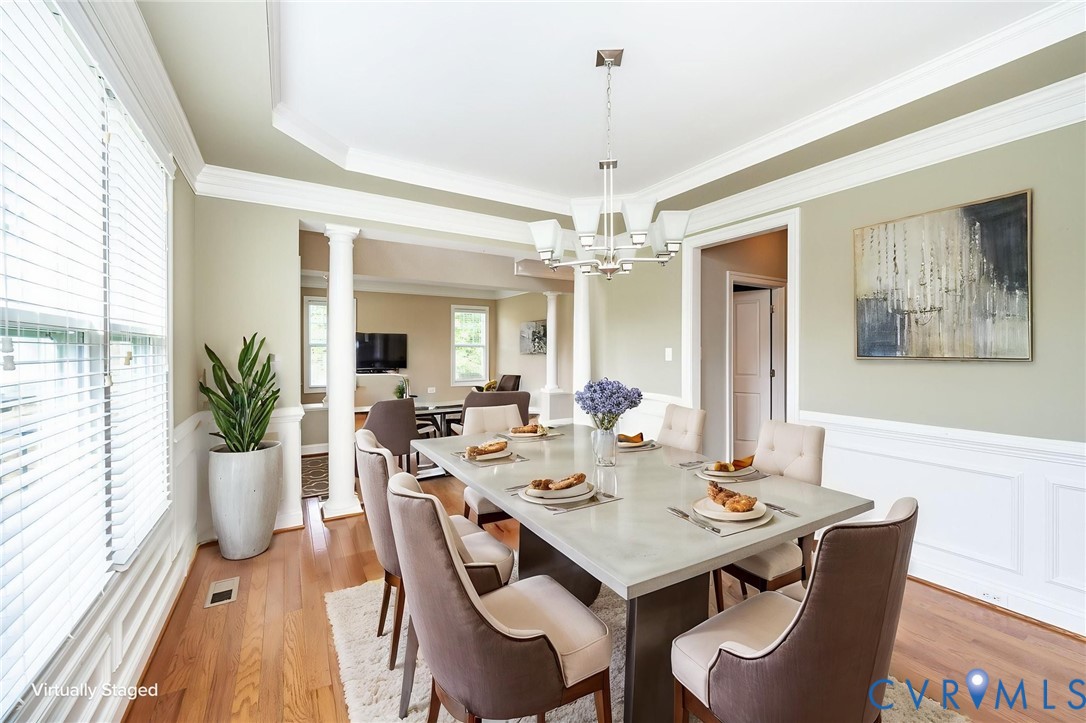 Dining room featuring wood-finish flooring, white wainscoting, a tray ceiling, structural columns, and a multi-arm chandelier