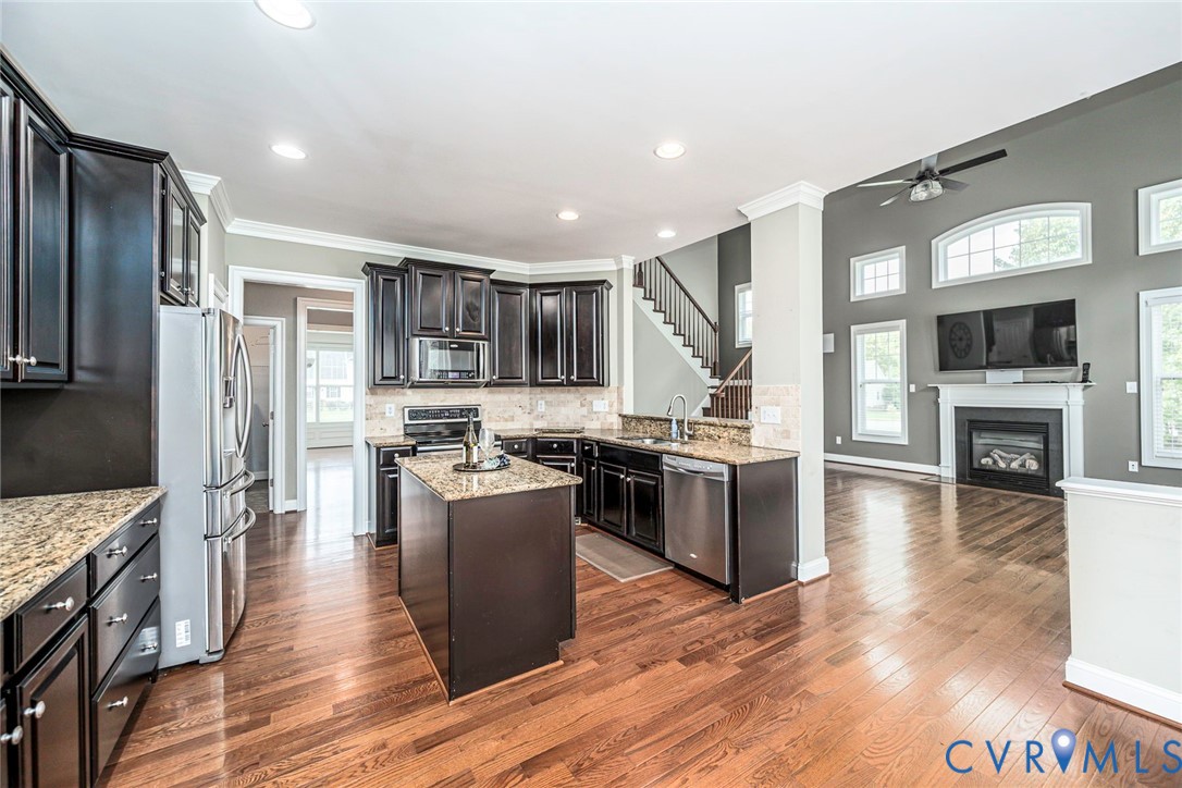 Kitchen featuring dark cabinetry, stone countertops, stainless steel appliances, wood-finish flooring, and a fireplace with a decorative mantel