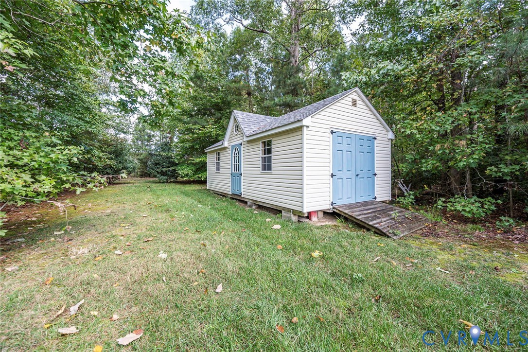 Detached outbuilding featuring vinyl siding, shingled gable roof, double door entry, and wooden accessibility ramp, situated on a grass-covered lot with mature trees