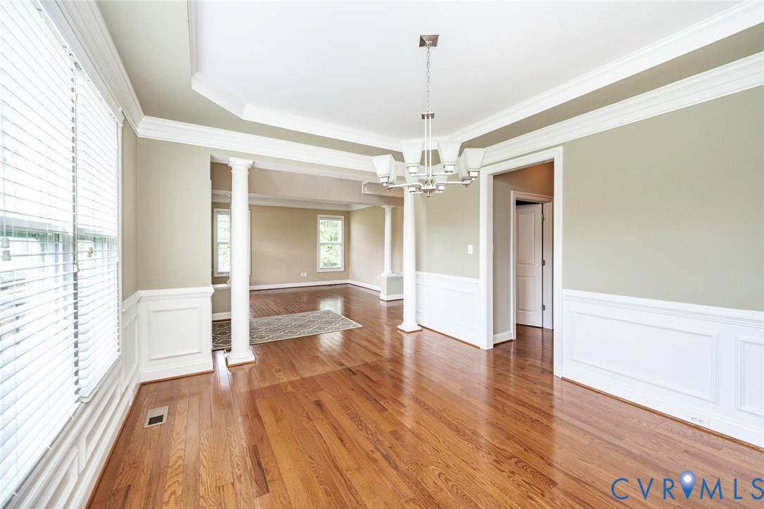 Wood-finish flooring, white wainscoting, decorative columns, tray ceiling, and multi-light chandelier
