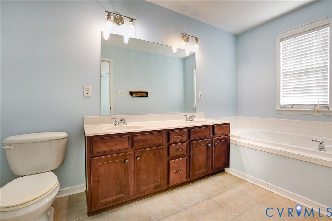 Bathroom featuring a wood-finish double vanity with an integrated countertop, large wall-mounted mirror, dual vanity lights, and a deep soaking tub with tile surround