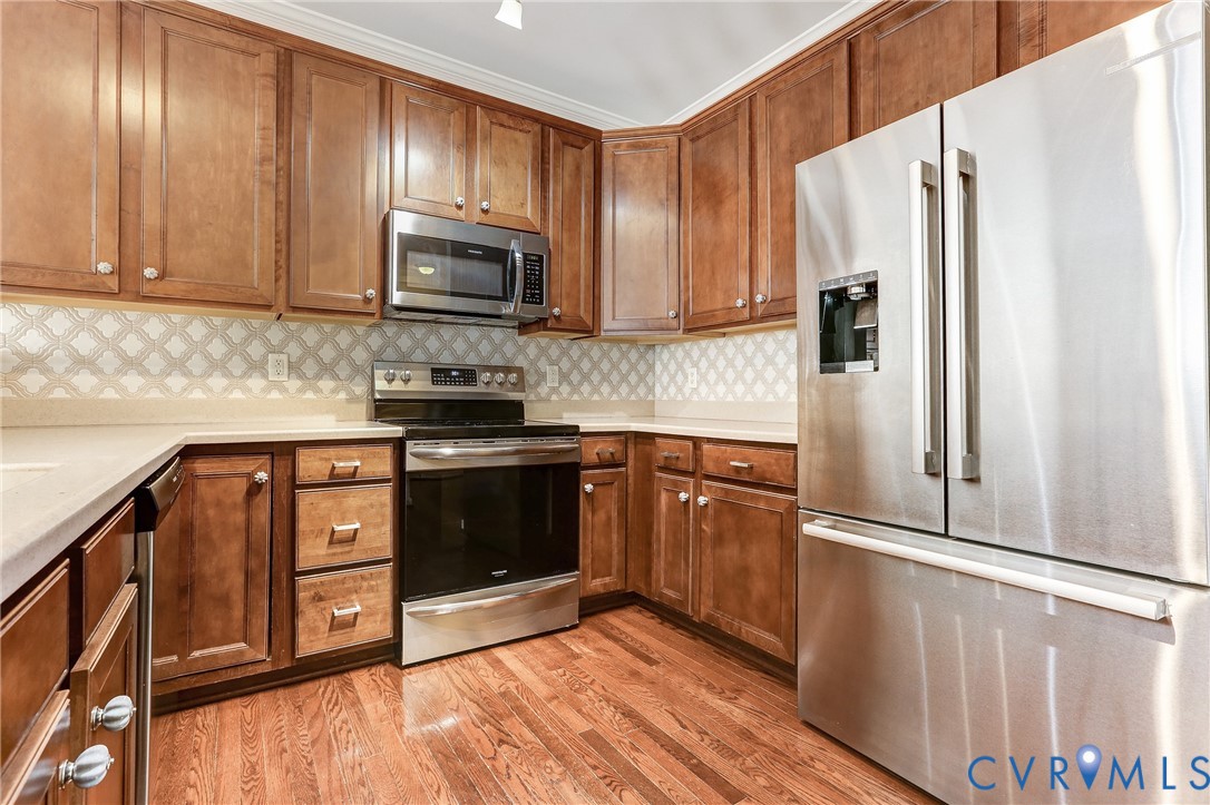 Kitchen featuring stained wood cabinetry, stainless steel appliances, solid surface countertops, a decorative patterned tile backsplash, and wood-finish flooring